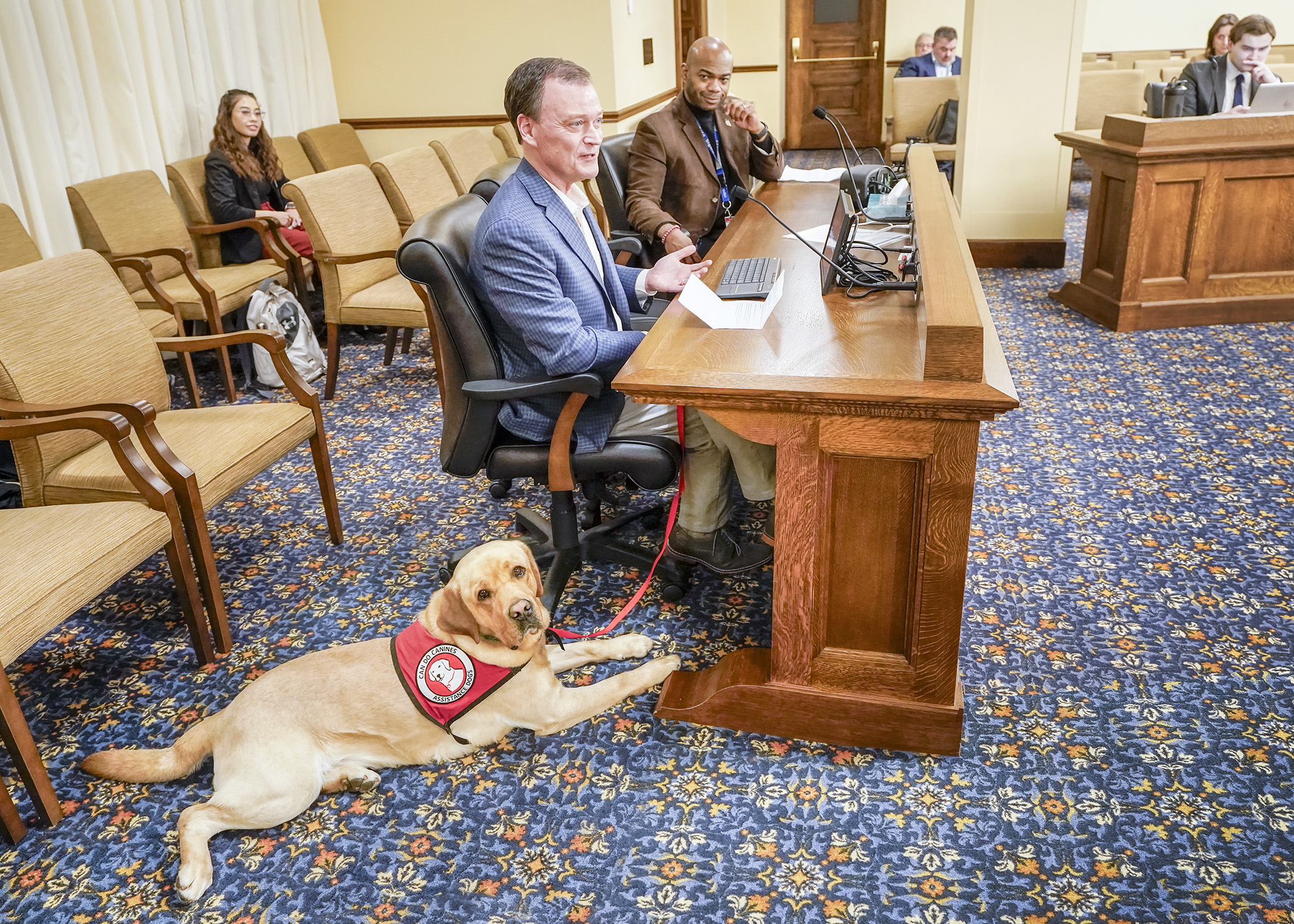 Jeff Johnson, executive director of Can Do Canines, testifies with Tucker, a 3-year-old service dog in training, during the House Housing Finance and Policy Committee Feb. 26. Johnson testified in support of a bill sponsored by Rep. Cedrick Frazier, seated right, that would modify service dog accommodations in housing such as rentals. (Photo by Andrew Von Bank)